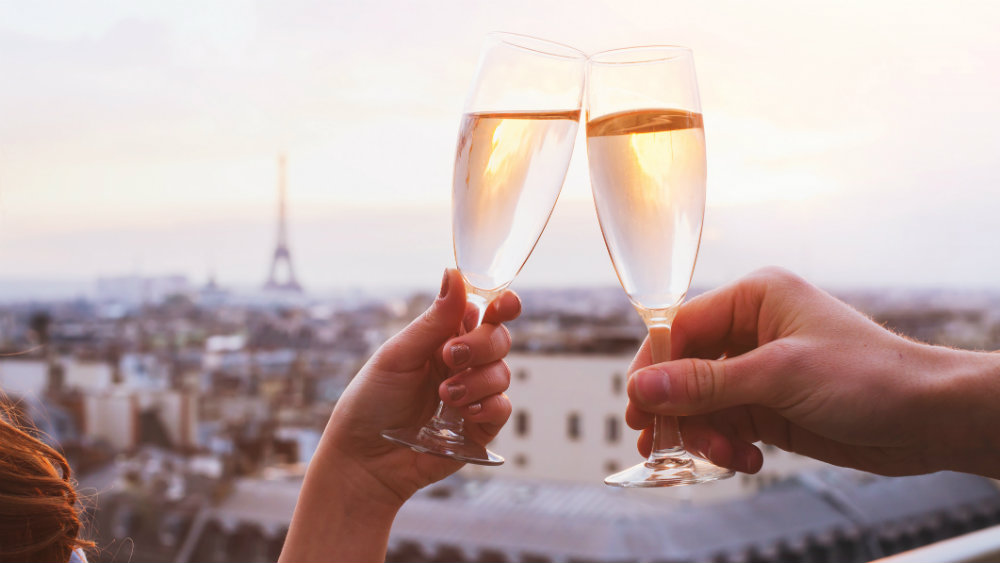 Two hands holding champagne glasses toasting each other with Paris in the background