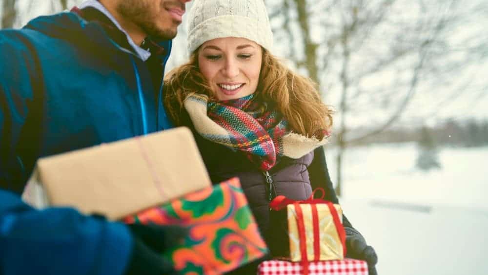 Smiling diverse couple holding Christmas presents while walking through a winter forest