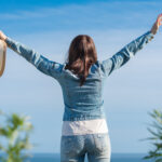 Female raising hands enjoying vacation, standing on background of blue cloudless sky.
