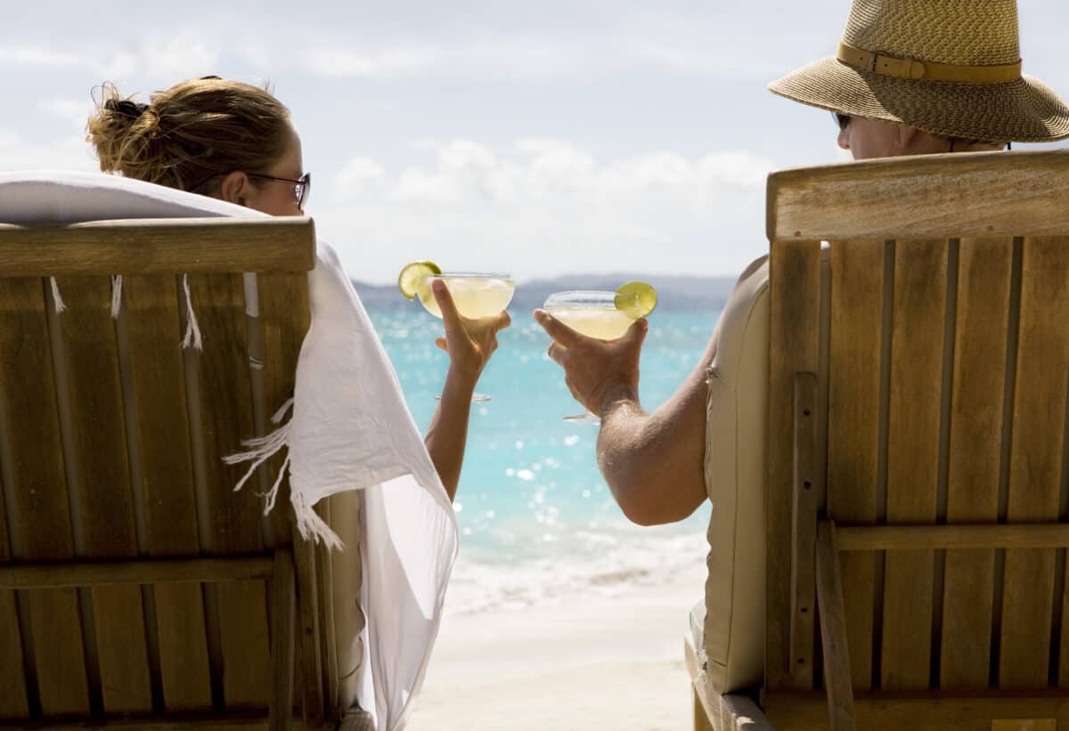 people sit in two wooden beach chairs facing the Caribbean ocean holding drinks and making a toast